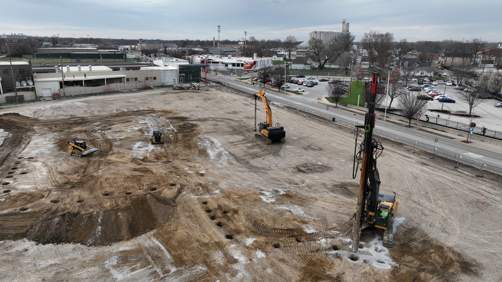 Aerial view of an active Pier Foundations ground-improvement site: a vibratory drilling rig and excavator installing stone columns on a prepared commercial pad with the city skyline behind.